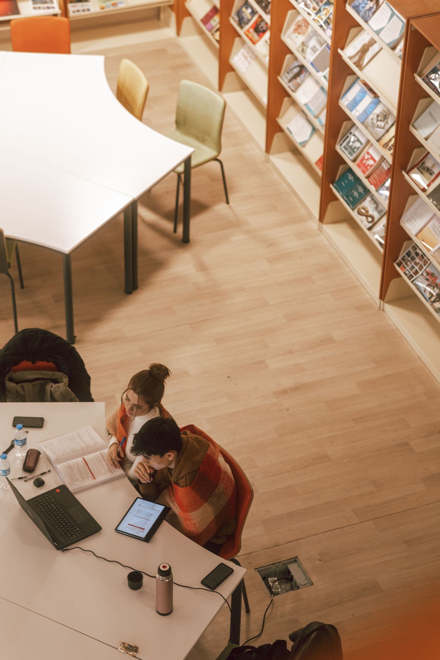 Vue de dessus : deux étudiants travaillent côte à côte à une table de bibliothèque, cahiers et ordinateur ouverts